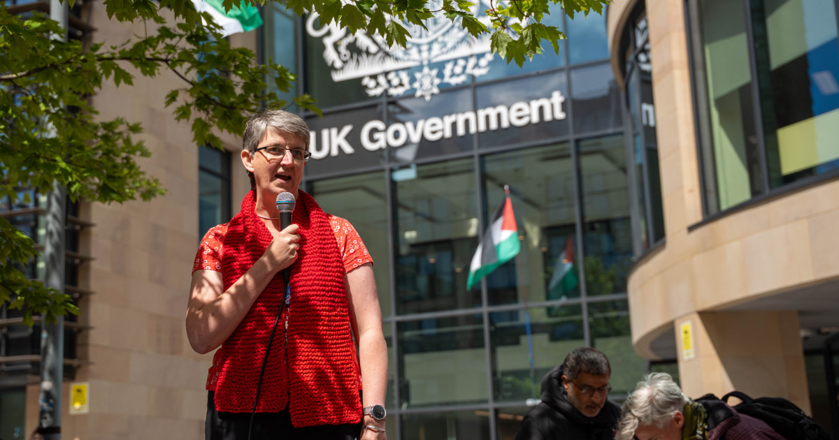 Maggie Chapman MSP speaks into a microphone to a crowd in front of the UK Government building. Palestinian flags can be seen in the background.