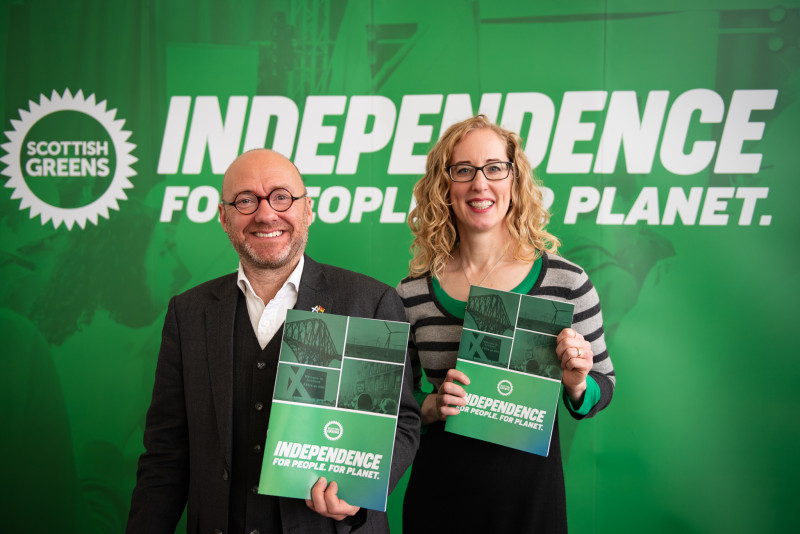Scottish Green Co-Leaders Patrick Harive MSP and Lorna Slater MSP standing in front of Independence For People, For Planet banner holding Independence policy papers.