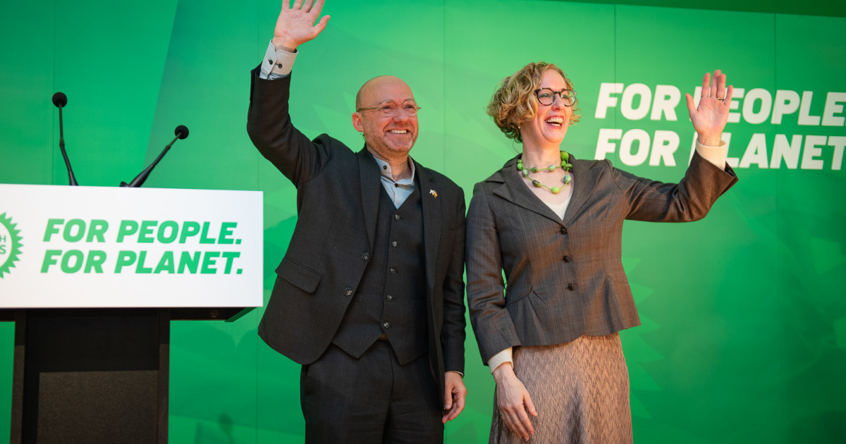 Scottish Green co-leaders Patrick Harvie MSP and Lorna Slater MSP stand on the stage of our 2023 Autumn Conference waving to the audience. Behind them words read 'For People. For Planet.'